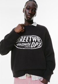 Sort strikket sweater med stort hvidt "STREETWR" logo, ribbede manchetter og kant. Båret over en lyseblåtern skjorte med krave. Lyserøde bukser synlige.
