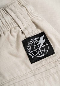 Beige fabric with a grid texture, featuring an elastic waistband and a black label with "REFLEX MEADOW" and a lightning bolt design.