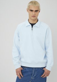 Jeune homme avec des cheveux blonds courts portant un pull léger bleu clair à col zippé et un jean bleu, se tenant les mains près des poches.