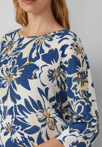Blouse florale en tissu blanc, bleu et marron. Présente une texture légère, un large col bateau, et des manches 3/4 avec un design plissé.