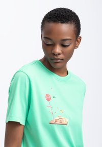 T-shirt in cotone verde menta con un design grafico che presenta un segnale di fermata e un'illustrazione di una trappola per topi sul davanti. Collo rotondo, maniche corte.