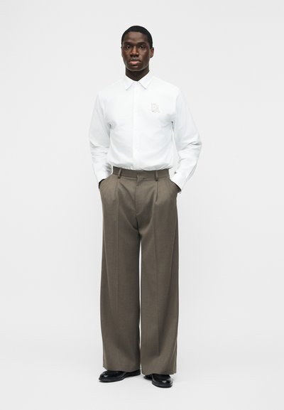 Άνδρας που φοράει λευκό πουκάμισο με μακριά μανίκια και κουμπιά με το λογότυπο "EA" και καφέ παντελόνι φαρδιάς γραμμής, στέκεται με τα χέρια στις τσέπες.