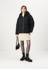 Zwarte pufferjack met diagonale stiksels, ritsluiting en capuchon. Gecombineerd met een crèmekleurige kabelgebreide jurk, zwarte schoenen en een kleine tas.