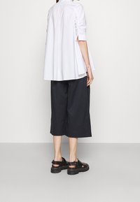 Camicia bianca con retro svasato e maniche arrotolate. Abbinata a pantaloni neri a tre quarti e sandali neri con suola spessa. Tessuto morbido, vestibilità comoda.