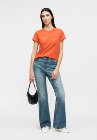 Γυναίκα που φορά πορτοκαλί μπλουζάκι και μπλε τζιν, κρατώντας μια μαύρη τσάντα, στέκεται μπροστά σε ένα απλό φωτεινό φόντο.