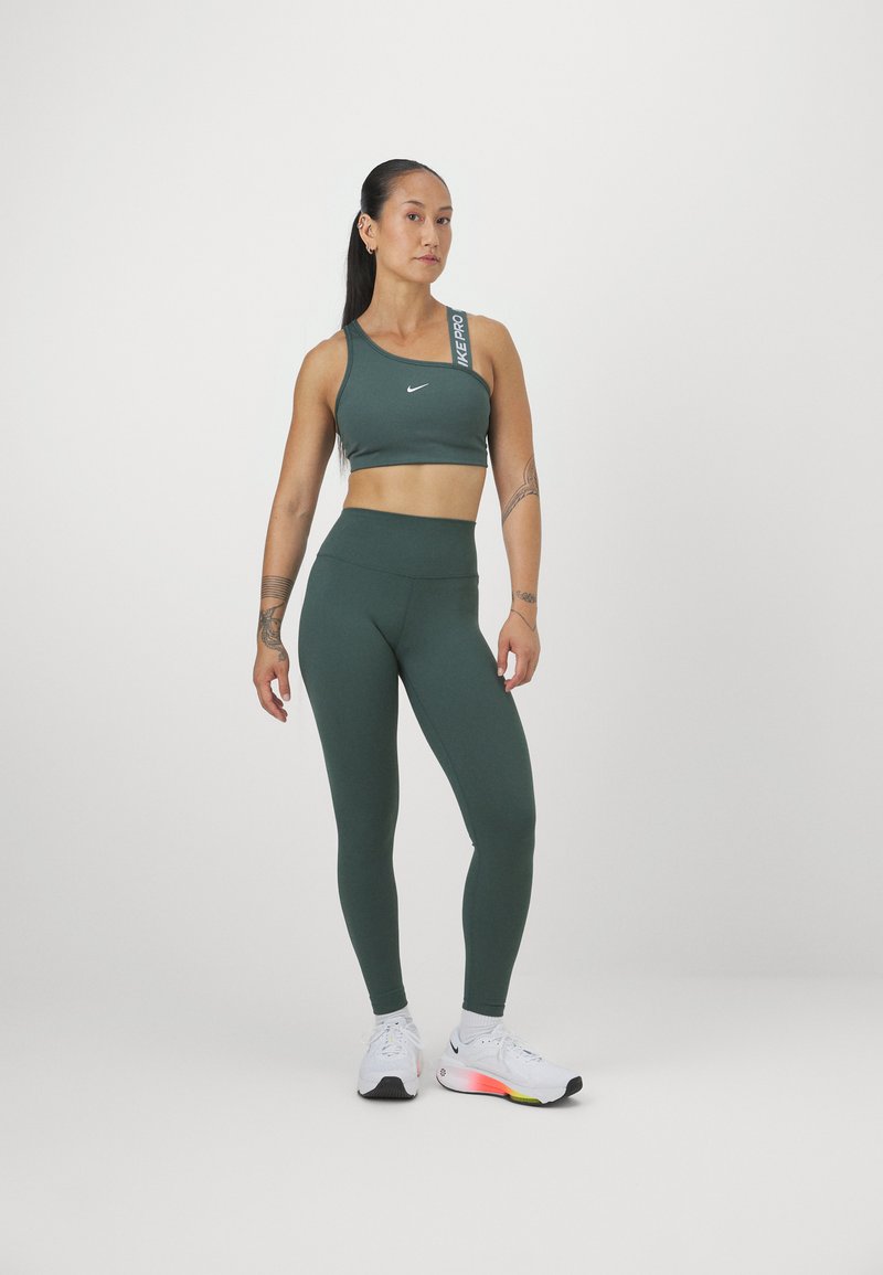 Donna in un reggiseno sportivo verde scuro a spalle scoperte e leggings abbinati, con scarpe da corsa bianche, contro uno sfondo neutro.