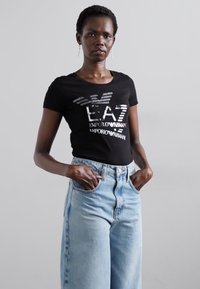 Zwarte t-shirt met metallic print met het logo "EA7 EMPORIO ARMANI"; gecombineerd met hoge taille lichtblauwe wijde jeans.