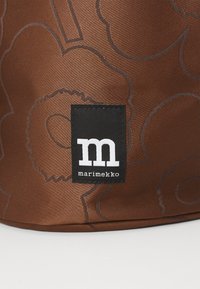 Sac en tissu marron avec un grand motif floral délimité en marron plus foncé. Il comprend une étiquette noire rectangulaire avec "m" et "marimekko."