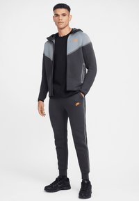 Pelēks sportisks krekls ar oranžām detaļām, plecu raksts, kopā ar atbilstošām sporta biksēm. Audums ir gluds un pieguļošs. Melni sporta apavi.