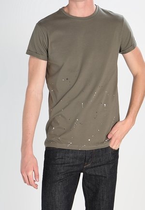 Mężczyzna ubrany w oliwkowo-zielony, krótki t-shirt z białymi plamkami farby i ciemnoniebieskie dżinsy, ręka w kieszeni, na jednolitym tle.