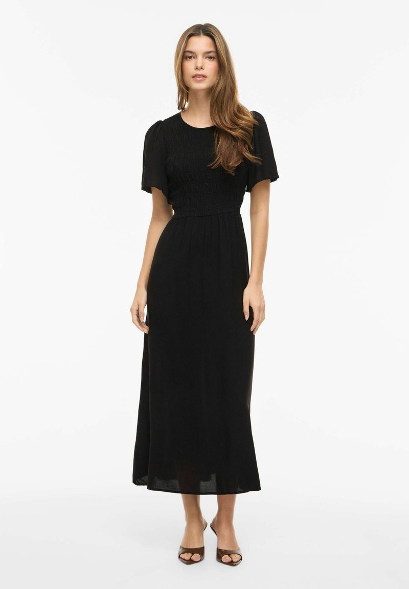 Mladá žena s dlhými vlasmi oblečená v čiernych midi šatách s krátkym rukávom a hnedých otvorených sandáloch na podpätku, stojaca na bielom pozadí.