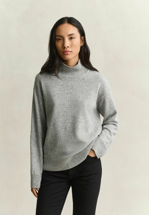 Vrouw met lang donker haar, gekleed in een grijze coltrui en zwarte broek, staand met één hand in de zak tegen een effen achtergrond.