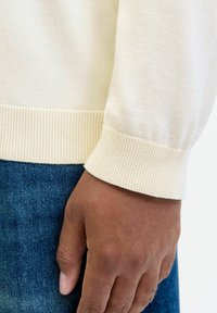 Crème-kleurige trui mouw met geribbelde manchetten, close-up van een hand tegen blauwe denimstof, waarbij de textuur en brei-details worden getoond.