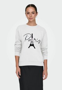 Mujer vestida con sudadera blanca con la Torre Eiffel y el texto "París", combinada con una falda negra, de pie contra un fondo liso.