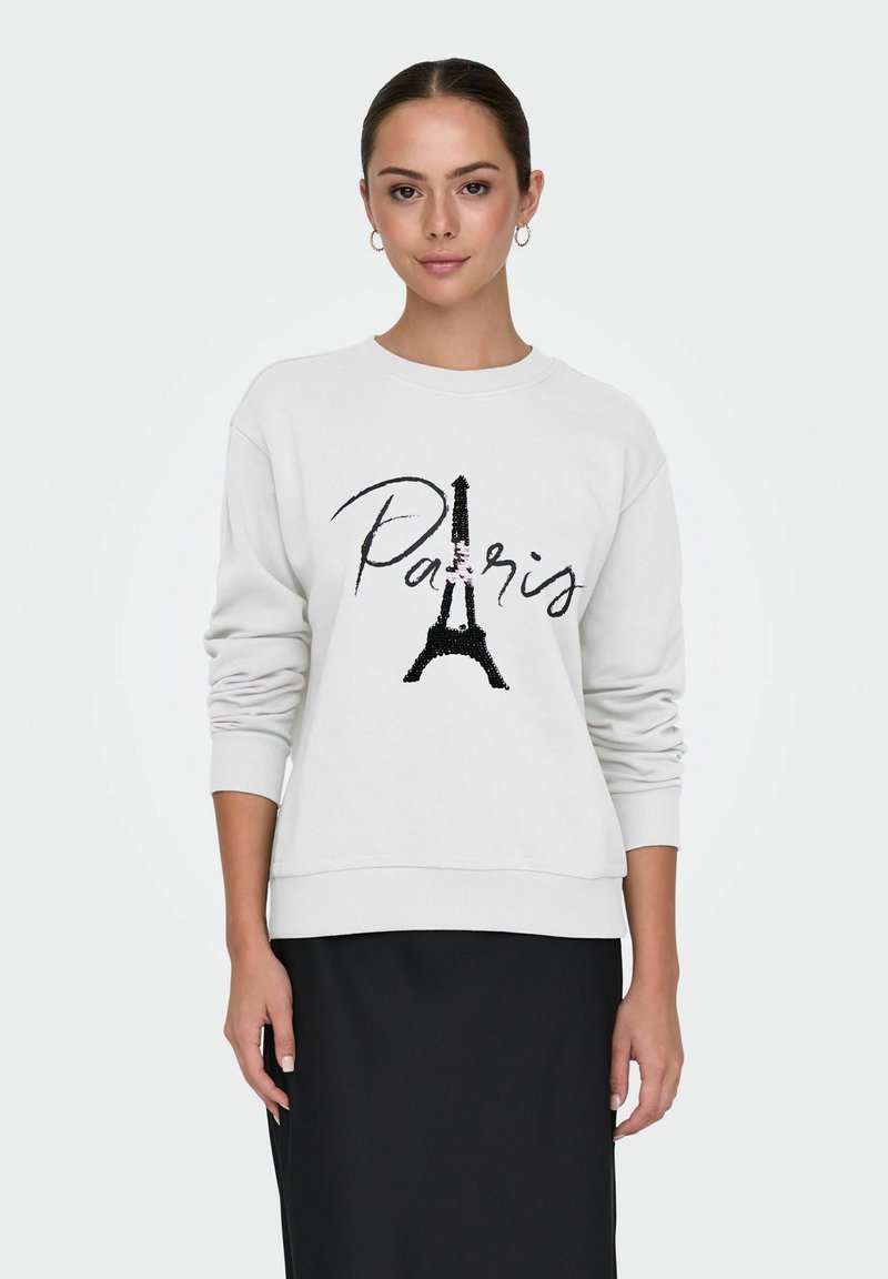 Mujer vestida con sudadera blanca con la Torre Eiffel y el texto "París", combinada con una falda negra, de pie contra un fondo liso.