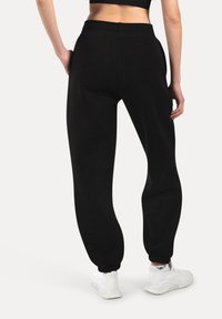 Henkilö, joka on pukeutunut mustiin jogging-housuihin ja valkoisiin tennareihin, kuvattuna takaa käsien roikkuessa rentoina sivuilla valkoista taustaa vasten.