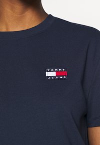 Detailní záběr na námořnickou modrou tričko s malou nášivkou Tommy Jeans na levé hrudi, nošené osobou se středně hnědou pletí.