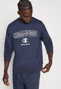 Μπλε ναυτικό φούτερ από βαμβάκι, με έντονα λευκά γράμματα που σχηματίζουν τη λέξη "CHAMPION" και λογότυπο, με ριμπ μανσέτες και τελείωμα.