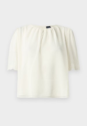 Blouse blanche à manches courtes avec encolure froncée et bords de manches brodés et festonnés, présentée sur un fond uni.