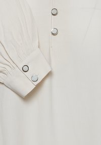 Blusa color crema realizzata in un morbido tessuto testurizzato, con maniche lunghe arricciate e tre bottoni a scatto in metallo lungo la patta frontale.