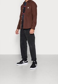 Camicia marrone con bottoni e dettagli sulla tasca, indossata sopra un maglione a righe blu e rosse, abbinata a pantaloni neri a sigaretta e sneakers nere.