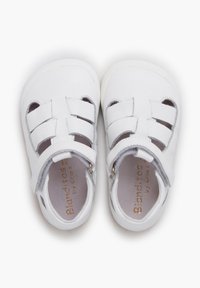 Weißer Ledersandal mit Überkreuz-Riemen-Design, offenen Seiten und verstellbaren Knöchelriemen. Glatte Textur und gepolsterter Innenraum. Goldene Markenkennzeichnung.
