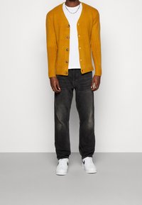 Hořčicově žlutý kardigan s pěti knoflíky, nošený přes bílé tričko. Kombinován s vybledlými černými džínami a bílými teniskami.