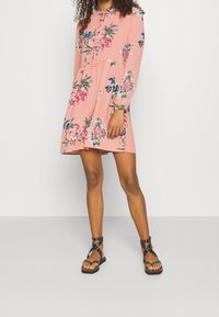 Vaaleanpunainen kukallinen mekko, jossa on pitkät hihat, nyörittäjä kaula-aukko ja virtaava hame. Mustat nauhalliset sandaalit, joissa on niittejä. Silkkinen kangas ja eläväiset kuviot.