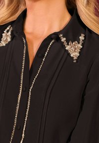 Svart blus med knäppning, dekorerad med pärl- och kristallpärldesigner på kragsprund och vertikal trim.