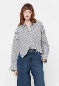 Junge Frau mit roten Haaren, die ein übergroßes weißes Hemd mit schwarzen vertikalen Streifen und hochgeschnittene, weite blaue Jeans trägt, steht vor einem weißen Hintergrund.