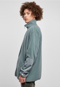 Fleece-pusero pehmeässä teal-värissä, jossa on korkea kaulus, harmaat yksityiskohdat hihoissa ja tasku; sisältää logotarkin hihassa.