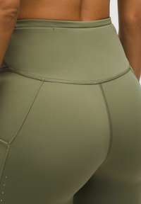 Hochtaillierte grüne Leggings aus glattem, dehnbarem Material; mit Nahtdetails und einer Seitentasche. Strukturierte Akzente entlang der Nähte.