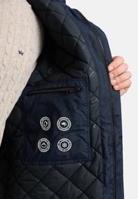 Intérieur d'une veste matelassée bleu marine avec une poche zippée et quatre écussons circulaires indiquant la résistance aux intempéries. La texture est lisse et brillante.
