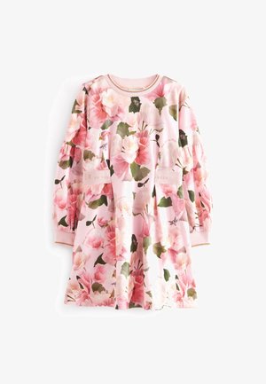 Enfant portant une robe fleurie rose à manches bouffantes et des baskets blanches, debout devant un fond uni clair.