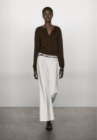 Cardigan marrone con bottoni, abbinato a pantaloni bianchi larghi. L'insieme presenta una cintura bianca in contrasto con dettagli metallici.