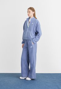 Fialový fleece teplákový súprava s bunda na zips a nohavicami s elastickým pásom. Obsahuje kontrastné šitie a malé vyšívané logo.