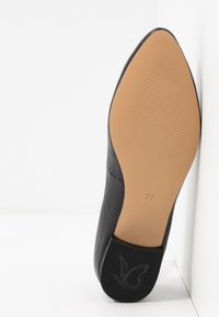 Chaussure femme noire à bout pointu inclinée sur le côté montrant une semelle lisse couleur fauve, taille 37 et petit logo papillon au talon.