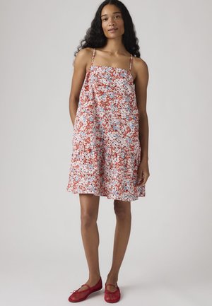 Žena s dlhými kučeravými vlasmi, oblečená v kvetinových šatách bez rukávov a červených balerínach, stojaca pred jednoduchým pozadím.