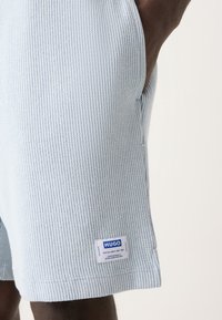 Shorts côtelés bleu clair avec poches latérales et une étiquette blanche portant l'inscription "HUGO" et des spécifications. Tissu texturé avec un motif vertical.