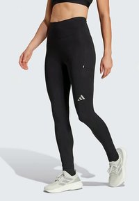 Zwarte high-waisted leggings met een gladde textuur, voorzien van een zijzak en het Adidas-logo in zilver op het linkeronderbeen.