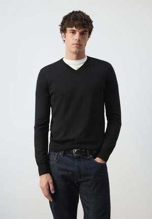 Joven con cabello rizado oscuro que lleva un suéter negro de cuello en V sobre una camisa blanca y vaqueros azul oscuro, de pie con una mano en el bolsillo.