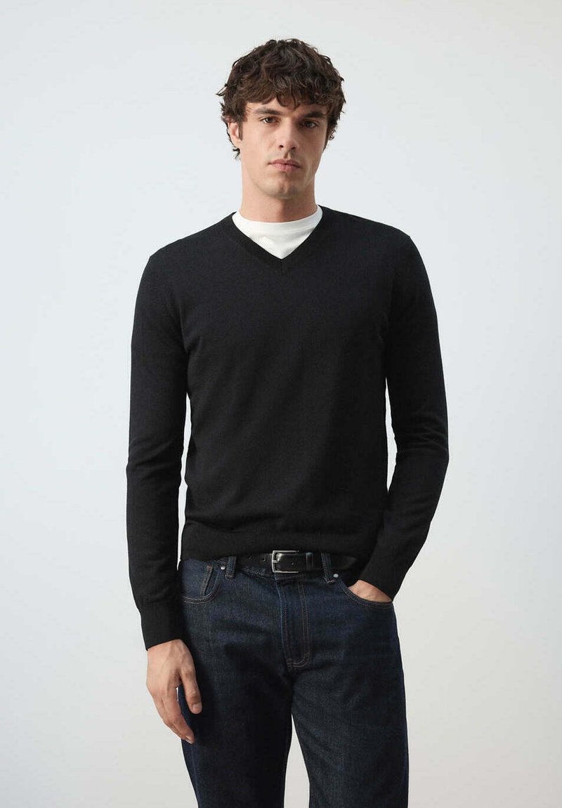 Jeune homme aux cheveux bouclés foncés portant un pull noir à col en V par-dessus une chemise blanche et un jean bleu foncé, debout avec une main dans la poche.