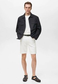 Giacca in tessuto scuro con due tasche sul petto, camicia bianca, pantaloni corti bianchi arrotolati e sandali neri. Tessuto liscio e design casual.