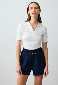 Witte geribbelde polo met korte mouwen, een V-hals en een gegolfde kraag, gecombineerd met marineblauwe shorts met elastische taille, zijzakken en gouden accenten.