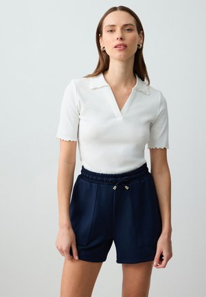 Polo bianco a righe con maniche corte, scollo a V e colletto scallop, abbinato a pantaloni corti blu navy con vita elastica, dotati di tasche laterali e dettagli dorati.
