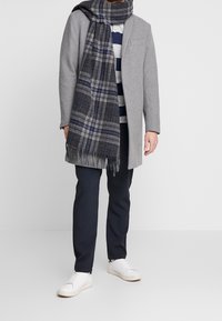 Cappotto di lana grigio chiaro, pantaloni scuri, sneakers bianche e una sciarpa a quadri grigio scuro con frange. Top a righe sotto. Sovrapposizione casual.