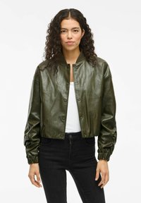 Grüne Leder-Bomberjacke mit cropped Design, versehen mit Druckknöpfen, geripptem Kragen und gerafften Bündchen, kombiniert mit schwarzen Jeans.