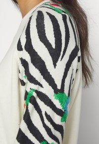 Gros plan d'une manche de pull blanc avec des rayures zèbre noires et de petits motifs floraux verts et bleus, portée par une personne aux longs cheveux bruns.