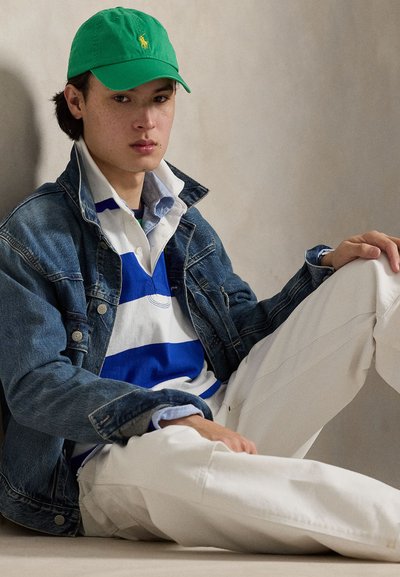 Denim jacket over blue and white striped polo, paired with white trousers, and a green cap featuring a yellow emblem. Simple and casual outfit.