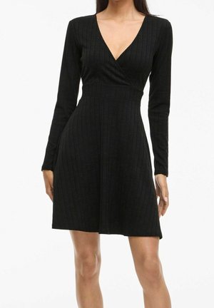 Vrouw draagt een zwarte, langhankelijke, geribbelde gebreide jurk met een V-hals en een getailleerde taille, staand tegen een witte achtergrond.